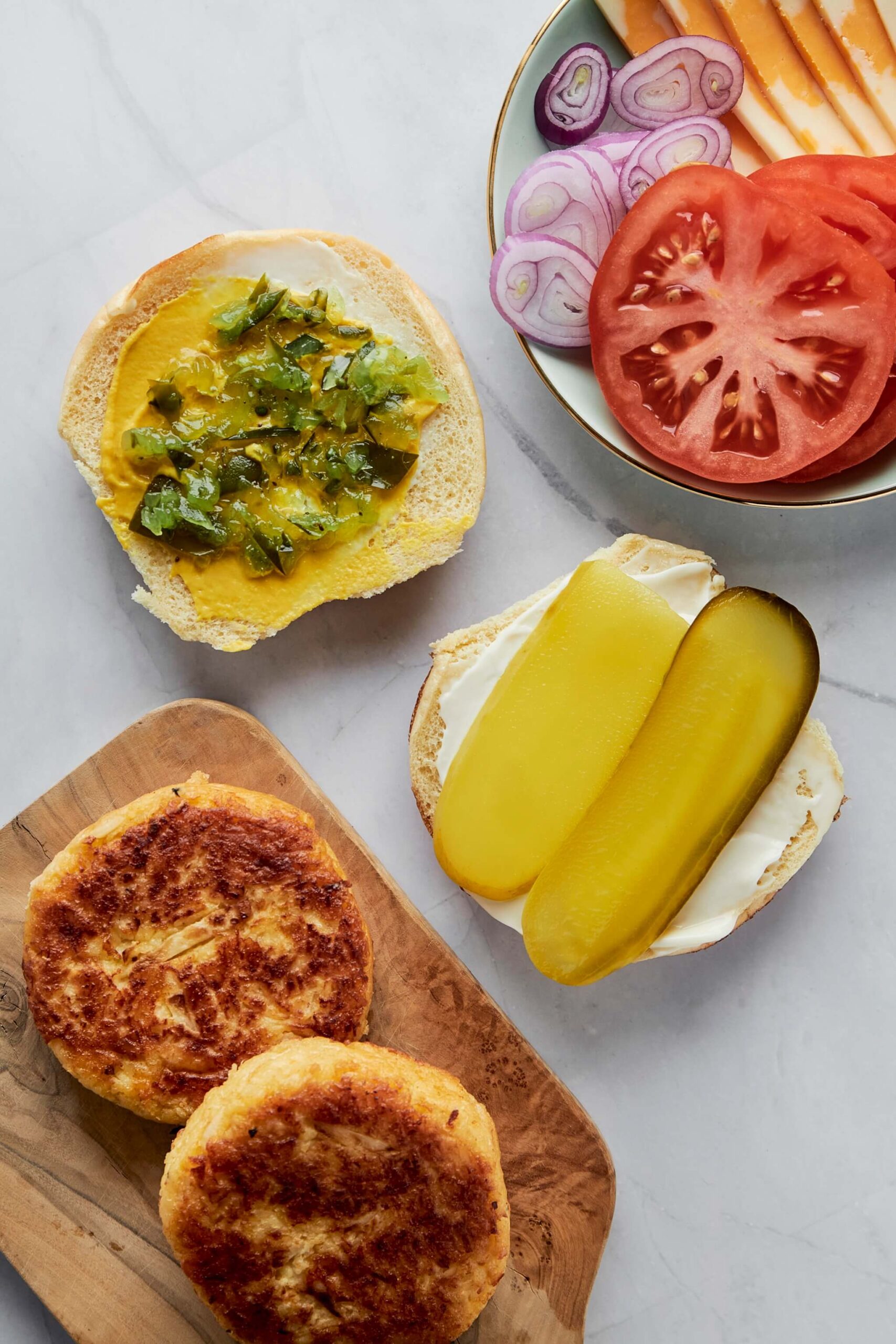 Assiette avec des tomates tranchées, des oignons rouges et des tranches de fromage, à côté de pains hamburger ouvert (la partie du haut, recouverte de moutarde et relish et la partie du bas recouverte de mayo et de pickles). En bas de l'image, deux galettes de burger au poisson