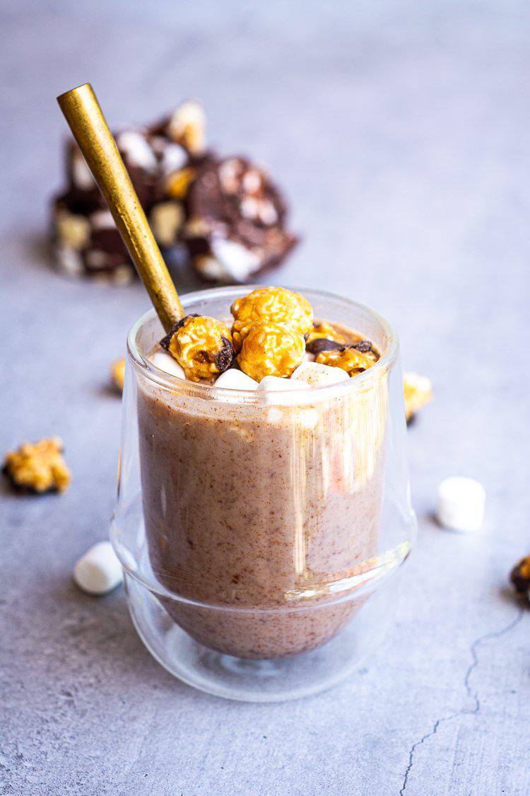 Verre transparent contenant le chocolat chaud vide-frigo avec ses garniture de popcorn et guimauves avec une cuillère dorée dans le verre